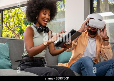 Amici diversi che testano il visore per la realtà virtuale condividendo il tablet sul divano grigio in salotto. Tecnologia, relax, modernità Foto Stock