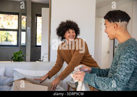 Amici afro-americani e asiatici che disimballano la scatola in movimento tenendo un asciugamano sul divano nel soggiorno. Accogliente, amicizia, casa, relax Foto Stock