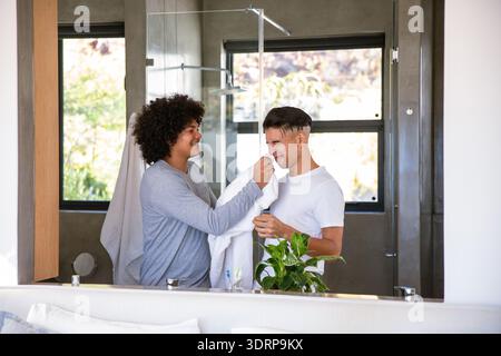 Due uomini in piedi nel moderno bagno di casa che puliscono la faccia con un asciugamano bianco davanti allo specchio. Romantico, intimo, moderno, benessere, cura del corpo, affetto, s Foto Stock