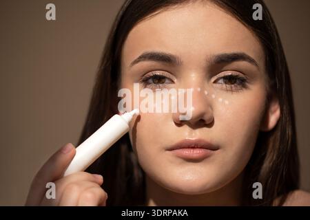 Giovane donna che applica prodotti per la cura della pelle sul viso, mostrando routine di bellezza con motivo tratteggiato sulla pelle, sottolineando l'autocura Foto Stock