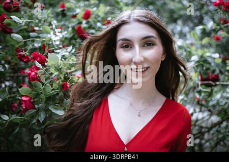 Giovane donna dai lunghi capelli marroni, con indosso una camicetta rossa, sorride gioiosamente in un lussureggiante giardino pieno di vivaci rose rosse, incarnano Foto Stock
