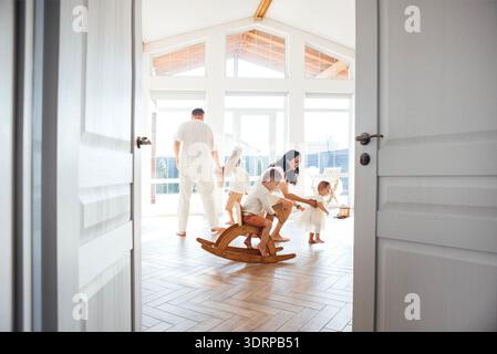 Famiglia che si diverte a divertirsi all'interno, bambini che giocano su un cavallo a dondolo di legno, genitori che si impegnano con i bambini, luminoso e spazioso Foto Stock