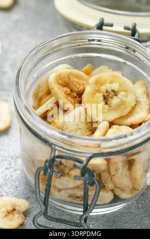Banane secche fatte in casa in un vaso di vetro e banane fresche. Fette di frutta fritte gialle. Spuntino dolce sano. Vegetarianismo Foto Stock