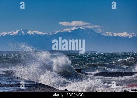 Onde potenti si infrangono contro la struttura costiera, mentre la cima innevata del Monte Olimpo si erge nettamente sullo sfondo. La scena viene evidenziata Foto Stock