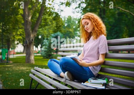 Un giovane legge un libro in un parco mentre è seduto su una panchina in un ambiente soleggiato Foto Stock
