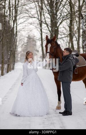 Sposa sorridente in un abito da sposa invernale bianco che tocca delicatamente un cavallo marrone su una strada innevata nella foresta, creando una romantica scena di matrimonio in campagna Foto Stock