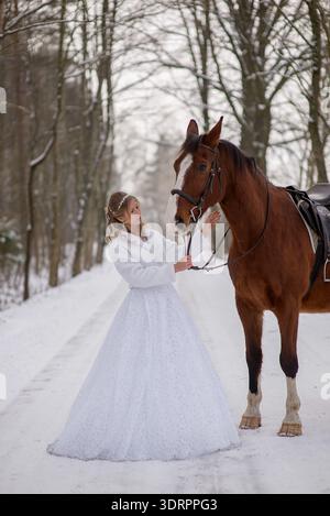 Sposa in piedi da sola con un cavallo sellato, tenendo le redini in un paesaggio boschivo innevato, mettendo in risalto eleganza, fiducia e un tranquillo matrimonio invernale Foto Stock
