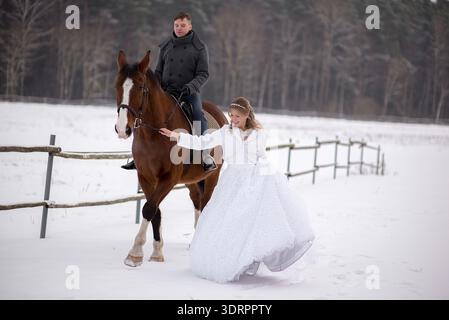 Scenario orizzontale per matrimoni invernali con una sposa sorridente in un abito bianco che cammina accanto a un cavallo marrone condotto dallo sposo attraverso un campo innevato, esprimendo Foto Stock