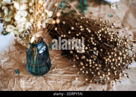 Il mazzo di lino essiccato e i fiori di ortensia si trovano in un vaso di vetro blu su carta marrone sgusciata. Bouquet di semi di lino, composizioni floreali, arredi naturali, sostenibilità Foto Stock