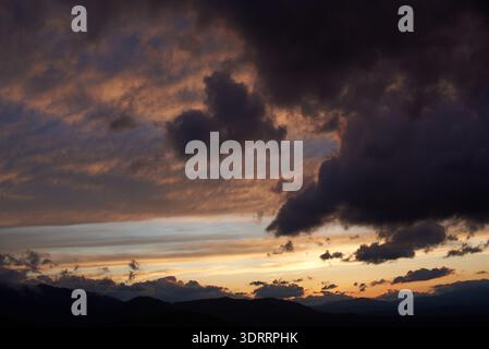 Spettacolare tramonto con nuvole scure e scoscese che proiettano ombre sul cielo. Horizon si illumina di calde tonalità arancio e rosa, contrastando con le silhouette delle montagne, creando un'atmosfera soffusa. Foto Stock