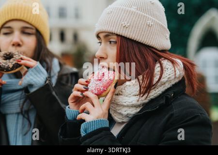 Due bambine in abito invernale mangiano ciambelle colorate all'aperto, sorridono e condividono dolcetti Foto Stock