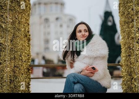 Una giovane donna con un cappotto di pelliccia bianca siede all'aperto, sorride e tiene in mano uno smartphone rosa. Foto Stock
