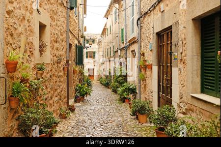 Stretta strada acciottolata a Valldemossa con case rustiche in pietra e persiane verdi. Tradizionale vicolo del villaggio di Maiorca decorato con piante in vaso Foto Stock