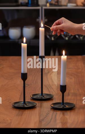 Primo piano di tre candeline accese. Una candela si accende con un fiammifero. Le candele sono bianche su un portacandele nero sulla parte superiore di Foto Stock