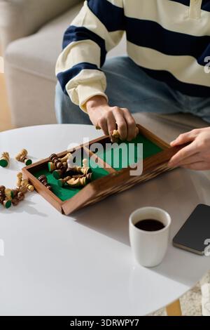 Un bel giovane organizza i pezzi degli scacchi su un tavolo accogliente mentre sorseggia il caffè. Foto Stock
