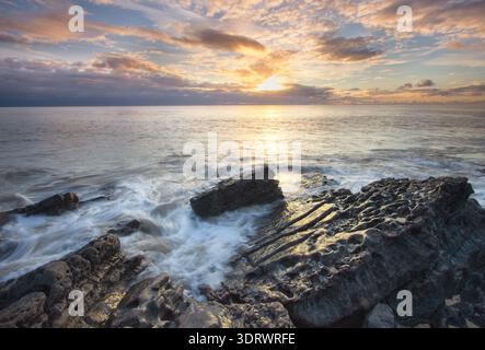 Costa rocciosa al tramonto con le onde che scivolano su sporgenze di pietra calcarea strutturata sulla Welsh Heritage Coast. Foto Stock
