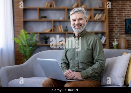 Uomo maturo con capelli grigi e barba sorridente per la fotocamera mentre si siede su un divano, utilizza un computer portatile e lavora in remoto in un ambiente confortevole da ufficio Foto Stock