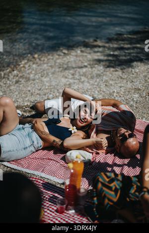 Gli amici si rilassano su una coperta a scacchi rossa e bianca vicino al fiume, godendosi una giornata estiva di sole Foto Stock