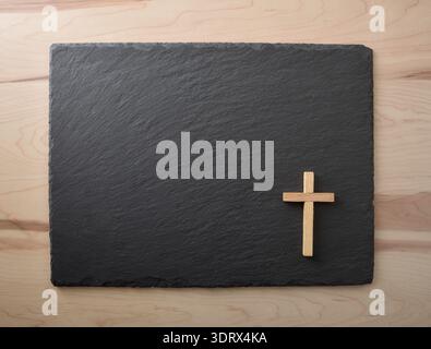 Una semplice croce di legno posta su una tavola di ardesia nera sopra un tavolo di legno chiaro. Composizione minimale con spazio di copia, simbolo del cristianesimo, della fede, a Foto Stock