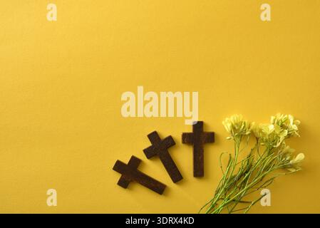 Tre croci rustiche in legno disposte su uno sfondo giallo brillante con delicati fiori gialli. Composizione piatta minima con spazio di copia, simboli Foto Stock
