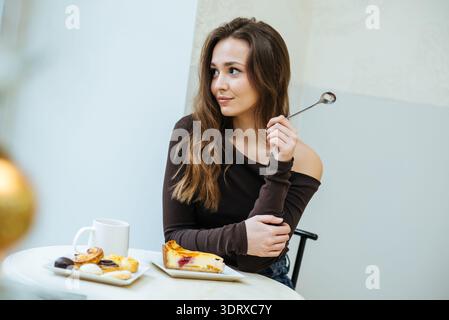 Una giovane donna siede ad un tavolino in un bar. Tiene in mano un cucchiaio e guarda con attenzione di lato. I piatti di dessert sono davanti a lei, insieme a.. Foto Stock