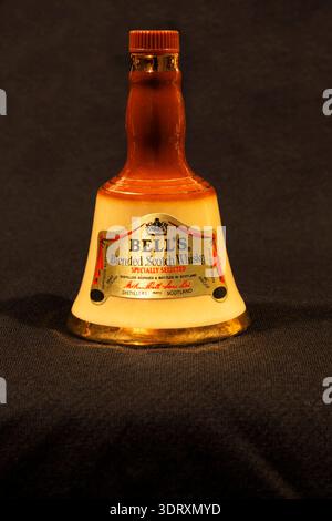 Primo piano della bottiglia di whisky scotch Blended in miniatura di Bell a forma di campana a mano con finiture dorate su sfondo grigio scuro in tessuto. Foto Stock