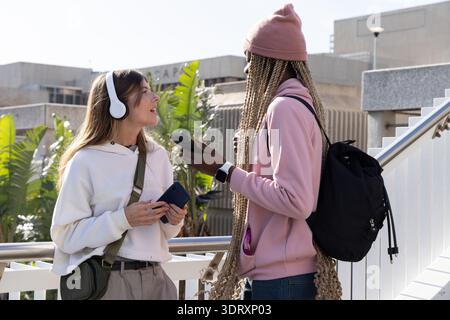 Diverse studentesse che chiacchierano sul passaggio pedonale del campus tenendo in mano smartphone e indossando felpe con cappuccio Foto Stock