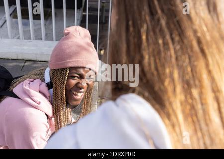 Ragazze adolescenti che chiacchierano su una sporgenza in cemento con ringhiere in metallo, una con berretto rosa Foto Stock