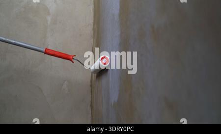 Rullo per pittura con impugnatura rossa che applica uno strato di primer bianco su una parete grezza appena intonacata in un angolo della stanza Foto Stock