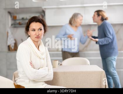 Donna frustrata seduta in cucina durante la lite tra il marito e la madre anziana. Foto Stock