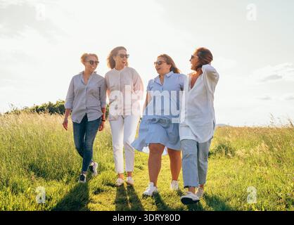 Allegra e variegata donna che si tiene mano nella mano camminando attraverso il prato verde per l'amicizia e la felicità delle donne con quattro splendidi ven Foto Stock