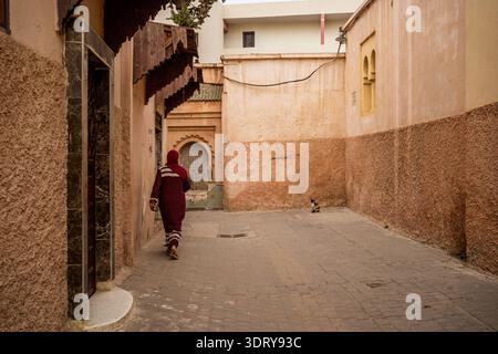 Una suggestiva vista posteriore di una donna che indossa una djellaba Bordeaux mentre cammina lungo una strada tranquilla nel cuore della Medina di Meknes, una lei mondiale dell'UNESCO Foto Stock