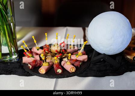 Vassoio di antipasti per catering con tartine assortite di involtini di carne su un tabellone nero, decorato su una tovaglia bianca accanto a una lampada a luna decorativa e un g Foto Stock