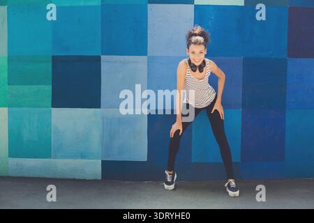 La giovane donna posa contro il muro del mosaico blu nell'area urbana durante il giorno con le cuffie accese Foto Stock