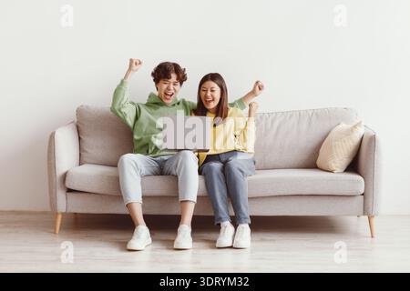 Giovane coppia asiatica con il laptop gesturing sì, celebrando il successo di affari in linea, seduta sul divano a casa, spazio copia Foto Stock