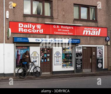 Una persona con una giacca marrone con cappuccio si stende con una bicicletta nera, guardando un telefono cellulare. Ciò si verifica al di fuori di G.B. Rodgers Off Licence Convenience sto Foto Stock