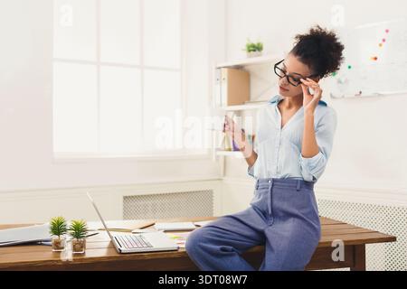 Donna seduta sulla scrivania in ufficio che tiene la tazza e indossa gli occhiali mentre guarda il laptop Foto Stock