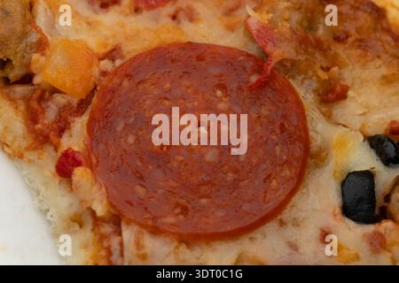 Primo piano di una fetta di pizza al peperoni con formaggio fuso, olive nere e peperoni, mostrando consistenza e dettagli saporiti Foto Stock