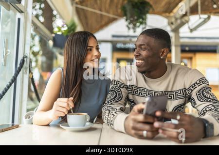 Coppie diverse che sorridono e interagiscono con gioia, condividendo un momento di relax insieme in un bar mentre guardano uno smartphone, simboleggiando la connessione e. Foto Stock
