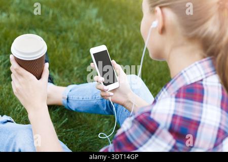Un giovane ascolta la musica mentre tiene in mano un caffè e utilizza il telefono sull'erba verde Foto Stock