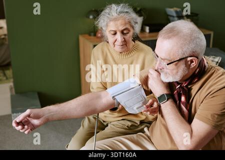 Uomo caucasico anziano che misura la pressione sanguigna con anziana donna caucasica che assiste Foto Stock