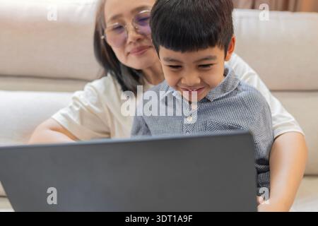 Allegra madre asiatica e simpatico figlio che ridono mentre si siede sul divano con un computer portatile a casa. Messa a fuoco selettiva. Foto Stock