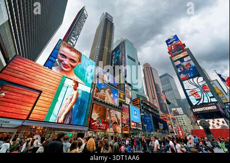 La gente cammina attraverso Times Square a Manhattan. I cartelloni luminosi mostrano pubblicità e immagini. Il cielo è nuvoloso sopra la strada trafficata. Foto Stock