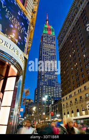 La gente cammina per la strada vicino all'Empire State Building, illuminato al crepuscolo di Manhattan, New York. La scena mostra la vita e le luci della città. Foto Stock