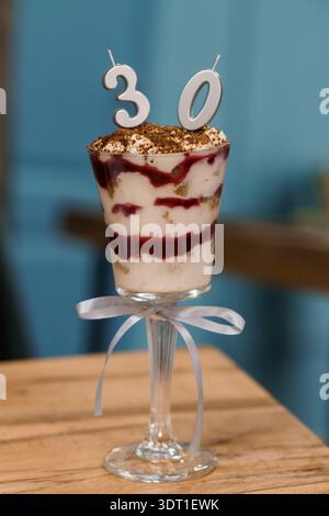 Elegante dessert del 30° compleanno in tazza di vetro con candele numerate, panna montata, bacche e polvere di cacao su tavolo di legno Foto Stock