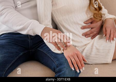 Un momento tenero tra una donna incinta e la sua compagna, entrambi con indosso bianco, toccando delicatamente l'un l'altro mani con una mano sulla pancia, trasmettendo Foto Stock