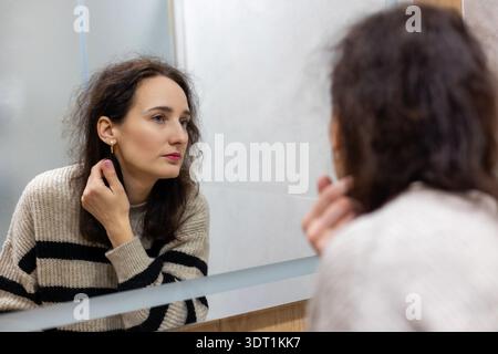 Donna di mezza età che guarda il viso nello specchio del bagno. Primo piano di una bruna di mezza età che guarda il suo riflesso nello specchio del bagno, calma Foto Stock