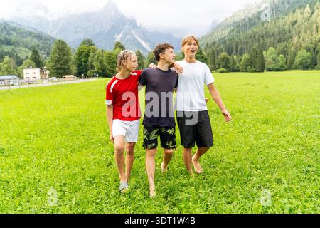 Un momento di gioia in famiglia durante una vacanza in montagna, con tre persone che camminano a piedi nudi su un lussureggiante prato verde con montagne panoramiche Foto Stock