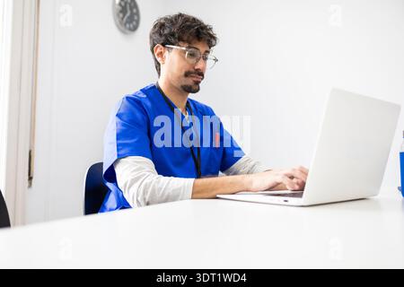 Un medico che indossa scrub blu è incentrato sull'utilizzo di un notebook alla scrivania di un ufficio moderno. L'ambiente professionale è completato da un orologio a parete A. Foto Stock
