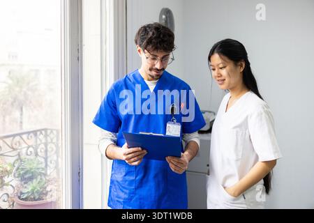 Un'équipe medica diversificata, tra cui un medico e un infermiere, collabora discutendo un piano di cura del paziente. Indossano scrub blu e bianchi, in piedi Foto Stock